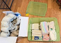 Overview of cardboard box with crib bumper and green tote containing baby textiles and accessories