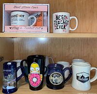 Shelf with boxed Mother mug, Best Teacher Ever mug, Chicago skyline mug, Homer Simpson mug, two blue stoneware mugs, three white mugs, Roadbucks mug