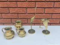All five brass items displayed against brick wall: two crane sculptures, three pitchers
