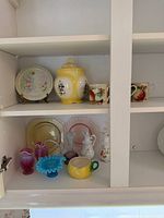 Cabinet shelf view showing porcelain floral plate, covered jar, fruit mugs, multiple glass and ceramic pieces