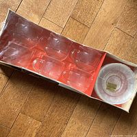 Factory box containing six clear glass cups and stack of six saucers, plastic wrap torn