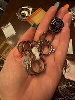 Hand holding eight assorted rings including signet and band styles, size tags visible