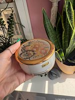 Full view of closed round porcelain box in hand showing size, gold rim, 24KT gold sticker