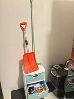 Red plastic snow shovel with aluminum shaft leaning against wall
