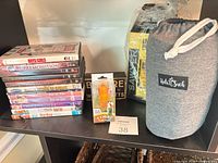 Shelf showing entire lot: DVD stack, corgi bottle stopper box, wood sign, Sawyer insect repellent pack, Kids N Such bag