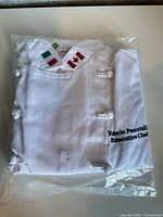 Front of folded chef jacket in plastic, showing flag patches and embroidery