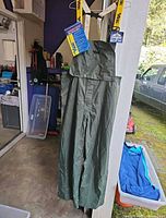 Full view of green waterproof bib pants hanging, showing yellow suspenders and attached tags