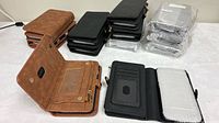Stacks of brown, black and gray wallet cases, two opened to show interior card slots, ID window, zipper pocket; plastic-wrapped stack of gray cases