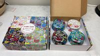 Two cardboard boxes, one closed lid shows floral collage design and product label, other open showing four tin candles