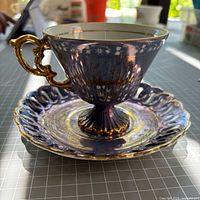 Side view of tea cup on saucer showing iridescent glaze, gold handle, pedestal base