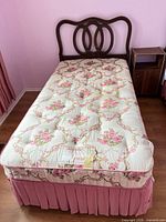 Twin bed showing wood headboard, floral mattress, pink skirt, nightstand partial view