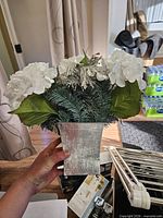 Front view of artificial arrangement held by hand showing silver metal vase and foliage