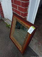 Full view of rectangular ornate gilded mirror leaning against brick wall