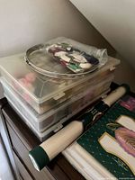 Stacked clear plastic organizers, metal hoops, floss skeins, fabric rolls and packaged Aida cloth visible