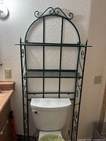 Full view of green wrought iron over-toilet shelf showing scroll sides, arched top, wire shelves, brass finials, positioned over toilet for scale