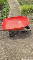 Side view of red plastic wheelbarrow showing tub, wooden handles, metal frame, pneumatic wheel