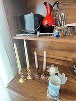 Shelf showing black ice bucket, red glass pitcher, metal bar tools, glass paperweight in wood dish, and various candle holders