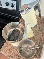 Mixer on countertop with large bowl, power cord inside, small bowl and beaters in foreground