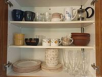 Full view of three shelves showing mugs, glasses, bowls, plates, French press