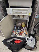 Open drawer and cupboard showing assorted tools, hardware, slide rule, vents, tape, bags