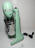 Left side view of mint green Hamilton Beach drink mixer with stainless cup attached and power cord visible