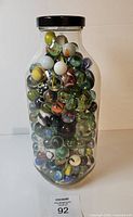Front view of tall clear jar packed with assorted vintage marbles, black lid visible