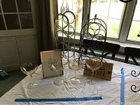 Photo showing two metal wine bottle holders, one holding 4 bottles and one 6, four peach tone wine glasses, and two framed wine quote decorations on a windowsill