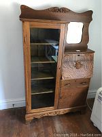 Front view of vintage oak secretary desk with glass door on left and drop-front desk on right, showing carved wood detail at top.