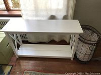 White wooden farm style sofa table placed in front of a window with curtain. Shows overall structure and condition of the table including wear and paint status.
