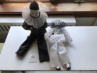 Pair of ceramic clown dolls seated on a white surface with a window background. One dressed in black polka dot outfit, the other in white polka dot outfit.