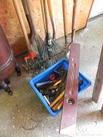 Photo showing multiple long-handled garden rakes and a red garden hoe leaning against a wall, with a blue plastic bin on the floor containing various hand tools including wooden-handled rubber mallets, chisels, and other woodworking tools.