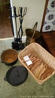 Photo showing basket filled with L.L.Bean fatwood sticks, two cast iron pots (one with lid), a rusted ash bucket, and a black metal fireplace tool set with four tools on a stand.