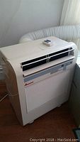 Full view of white portable Amana air conditioning unit with exhaust hose attached and remote control resting on top.