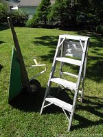 Metal wheel barrow with wooden handles and green bin on grass next to aluminum step ladder