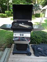 Full frontal photo of black PGS gas grill with lid open, side shelves extended, grilling tools inside, propane tank on lower shelf, and grill cover beside unit on deck.
