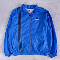 Front view of blue adidas windbreaker showing black triple stripe, full zip, pockets, elastic cuffs and hem