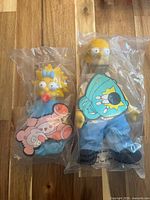 Front view of Homer Simpson and Maggie Simpson plush dolls in sealed plastic bags with cardboard tags visible