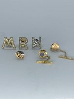All six pins and tacks arranged: three rhinestone letters M R N, FH monogram pin, two tie tacks with chains