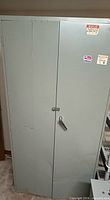 Front view of gray metal storage cabinet showing double doors, handle with lock hasp, gray painted surface, dents and scratches