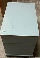 Front angled view of light gray two-drawer metal mobile filing cabinet showing lock and surface wear