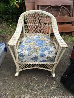 White painted wicker rocking chair with floral cushion seen front facing outdoors on gravel.