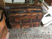 Front view of the antique dome top trunk showing wooden slats and original metal hardware, with visible wear and signs of aging.