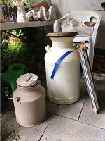 Pair of painted old milk cans with lids and metal expandable screens next to them on a concrete floor with chair and plants visible.