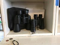 Two pairs of binoculars side by side with a black carrying case behind them on a shelf; one larger Mercury model 1111, one smaller textured pair