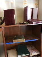 Four photo albums on wooden cabinet shelves: two maroon albums on top shelf (one open), a green album in middle shelf with old worn cover, and another green album on bottom shelf.