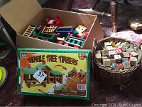 Wicker basket filled with assorted wooden letter and number blocks and toys, cardboard box with additional wooden toy parts including red and green painted components and wheels seen in background