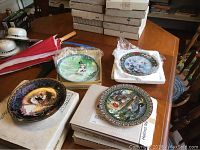 Four Bradford Exchange collector plates on a wooden table, some in their original boxes and one wrapped in protective plastic.