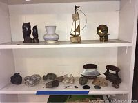 Shelf with two wooden carved owls, white ceramic vase, metal sailboat on marble base, various rock specimens, two vintage metal irons, and a brass lamp.