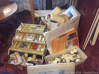 Photo showing assortment of art supplies including paint jars, paintbrushes in organized metal tray, wooden thread spools, trays and packing materials