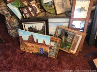 Collection of assorted framed paintings and mirrors placed on floor against furniture, showing various art styles including landscapes, botanical prints, and decorative mirrors.